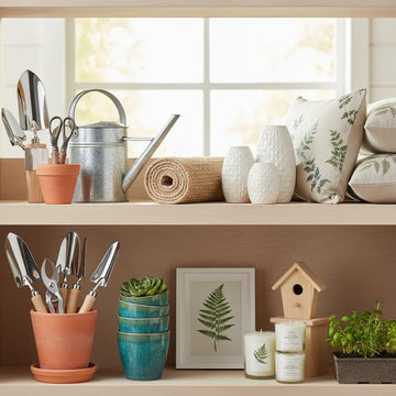 Home and garden products including gardening tools, watering can, pots, indoor plants, throw pillows, candles, and vases on wooden shelves.